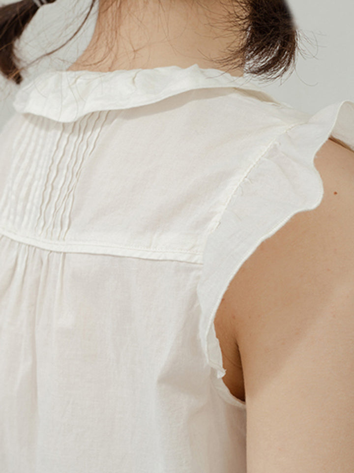 モデルがフランス少女プリーツ付きフリルスリーブレス白コットンシャツを着用し、エレガントなスタイルを披露。
