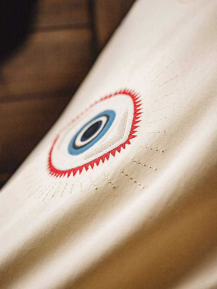 Tシャツ前面に施された「守護の眼」刺繍のクローズアップ。使用された7色の糸と複数の刺繍技術が生み出す複雑さを強調。