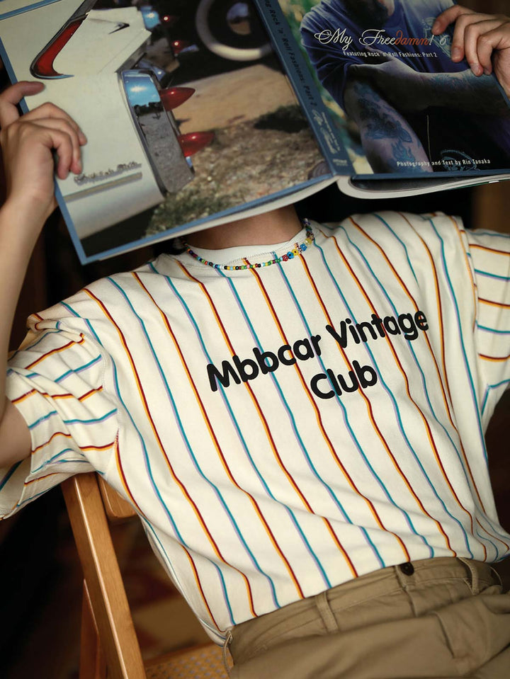 「Mbbcar Vintage Club」プリント付きの鮮やかなストライプTシャツを着用するモデル。ベージュの地に配された色彩がレトロな雰囲気を強調。