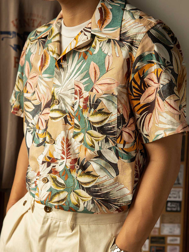 モデルがハワイアンプリント半袖シャツを着用し、無地のカジュアルパンツとシンプルなサンダルを合わせたスタイルでポーズ。明るく夏らしいカジュアルスタイルを演出。