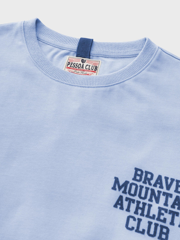 マカロンカラーのTシャツの「BRAVE MOUNTAIN ATHLETIC CLUB」フロッキープリントのクローズアップ、テクスチャとディテールを強調。